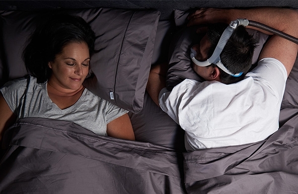Pärchen liegt im Bett, Mann liegt mit CPAP Maske auf dem Bauch und schläft