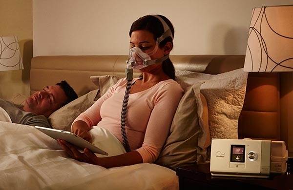 CPAP Anwenderin sitzt mit CPAP Maske im Bett mit Tablet in der Hand, AirCurve Gerät steht auf dem Nachttisch