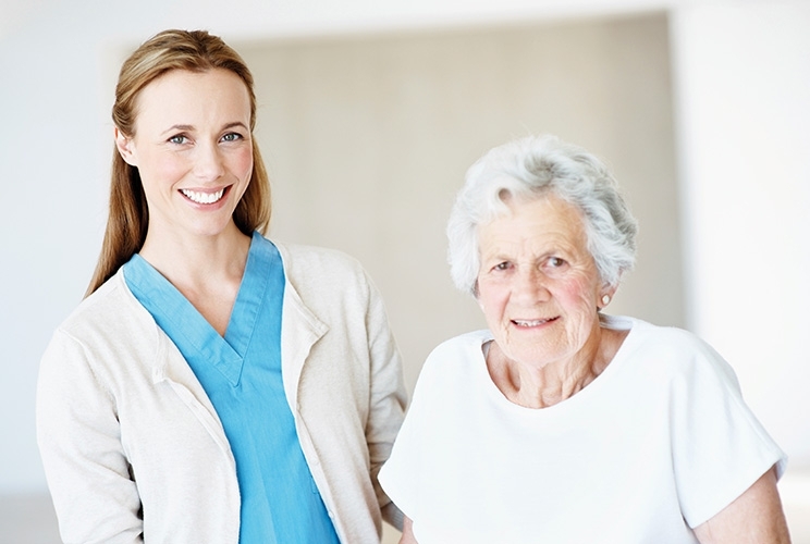 Pflegerin und ältere Patientin stehen nebeneinander, blicken in Kamera und lächeln