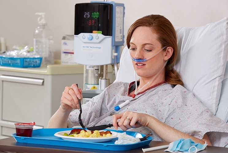 Patienten wird mit Vapotherm durch Sauerstoffbrille beatmet, sitzt aufrecht im Krankenhausbett und isst eine Mahlzeit