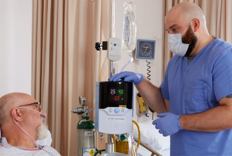 Pfleger steht bei Patient am Bett, der mit Vapotherm über eine Sauerstoffbrille beatmet wird und spricht mit ihm