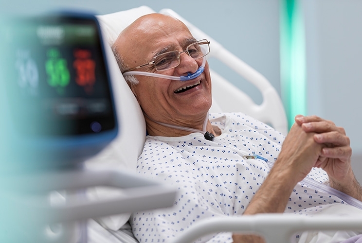 Älterer Mann sitzt in Krankenhausbett mit Nasenkanüle, wird mit HVT 2.0 beatmet blickt in die Ferne und lächelt