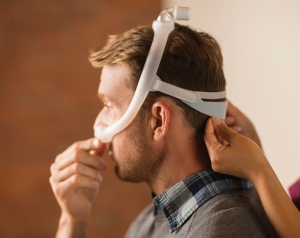 Ein Mann hält seine DreamMaske fest, eine Pflegerin hilft ihm, die Haltebänder über den Hinterkopf zu ziehen