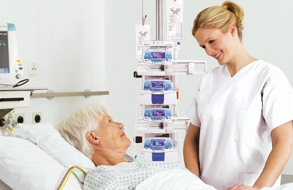 Eine Pflegerin lächelt eine Patientin an, im Hintergrund sieht man mehrere Agilia SP TIVA-Geräte im Haltesystem gestapelt