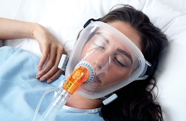 Patientin liegt im Krankenbett mit den Augen geschlossen und wird mit Vollgesichtsmaske beatmet