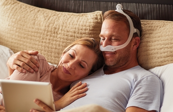 Ein Paar liegt kuschelnd im Bett, der Mann hält ein Tablet und trägt die DreamWear Maske
