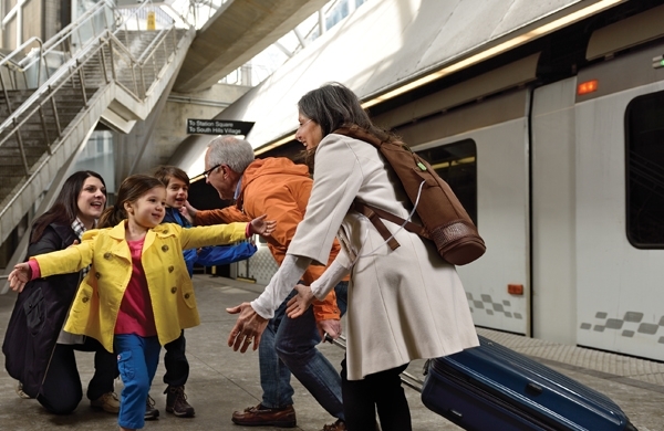 Enkelkinder begrüßen freudig ihre Großeltern am Bahnsteig, die Großmutter trägt den SimplyGO Mini im Rucksack am Rücken