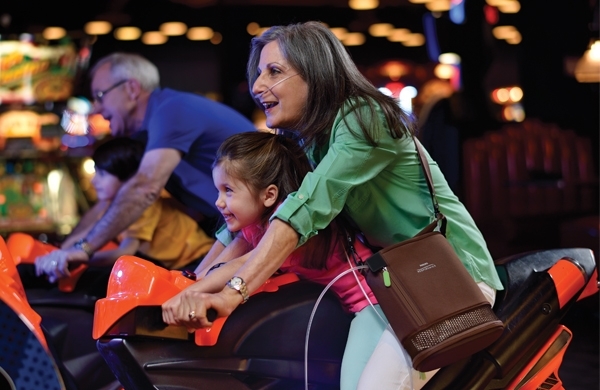 Großmutter vergnügt sich mit ihrer kleinen Enkeltochter in der Spielhalle bei einem Motorrad-Spiel, sie trägt den SimplyGO Mini in der Umhängetasche