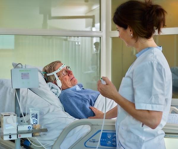 Patient liegt im Krankenhausbett an DeltaScan angeschlossen und hat die Augen geschlossen, Krankenschwester führt Delir Messung durch