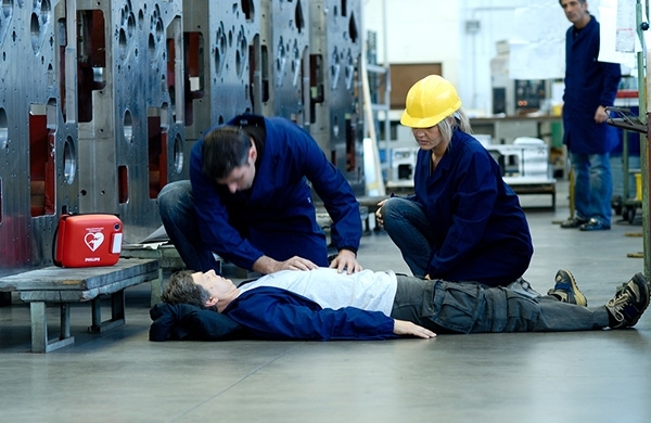 Arbeiter liegt bewusstlos am Boden bei seinem Spind, Kollege und Kollegin knien bei ihm, der Defibrillator steht auf einer Bank daneben