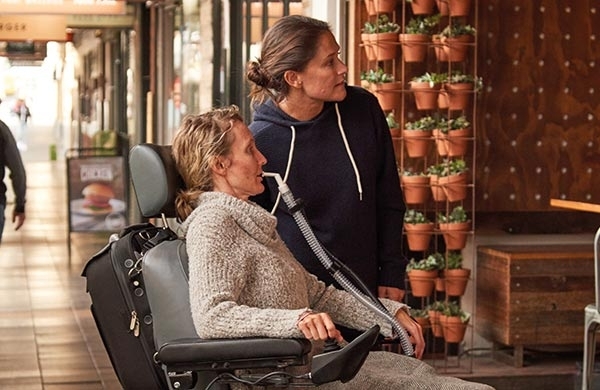 Patientin sitzt im Rollstuhl und ist mit ihrer Pflegerin auf einem Markt, wird mit Astral über ein Mundstück beatmet