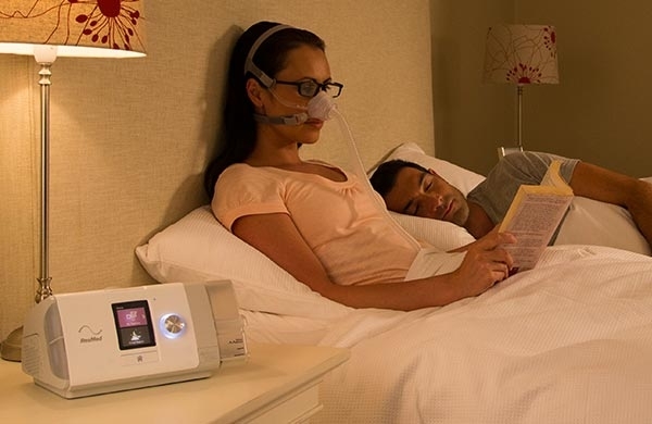 CPAP Anwenderin sitzt mit CPAP Maske im Bett mit Buch in der Hand, AirCurve Gerät steht auf dem Nachttisch