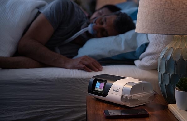 AirSense steht auf dem Nachttisch und CPAP Anwender schläft mit CPAP Maske im Bett