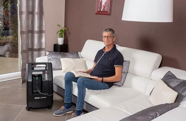 Mann mit Sauerstoffbrille sitzt im Wohnzimmer auf der Couch, Sauerstoffkonzentrator Compact 525 steht neben ihm auf dem Boden