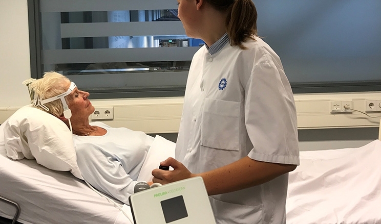 Patientin liegt im Krankenhaus im Bett, Pflegerin lehnt am Bett und führt DeltaScan Messung bei Patientin durch
