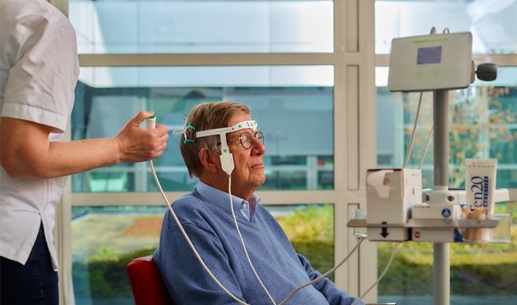 Patient sitzt auf Stuhl im Krankenzimmer, Pflegerin führt bei ihm Delir-Messung mit dem DeltaScan durch