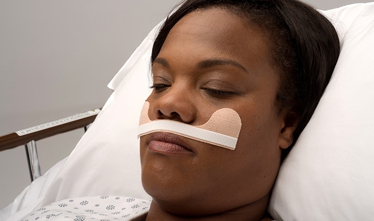 Patientin liegt mit geschlossenen Augen in Krankenhausbett mit Klebestreifen zwischen Mund und Nase
